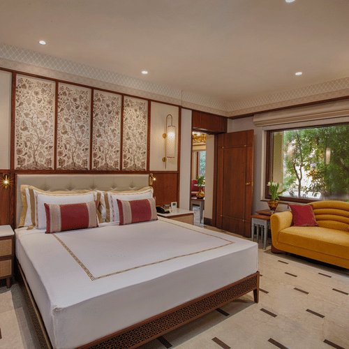 A bedroom with a king size bed, elegant floral touches, a cheerful yellow sofa, and a large window offering serene views of lush greenery | Heritage Village Resorts & Spa, Manesar.