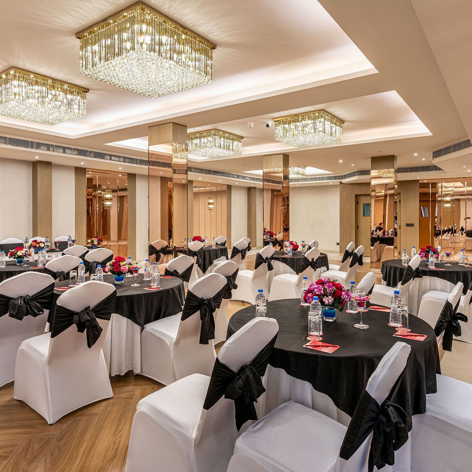 A banquet hall with tables and chairs arranged for dining, adorned with black tablecloths and chair sashes. It features a decorative ceiling with hanging lights and mirrored panels on the walls - Lords Eco Inn Dehradun.
