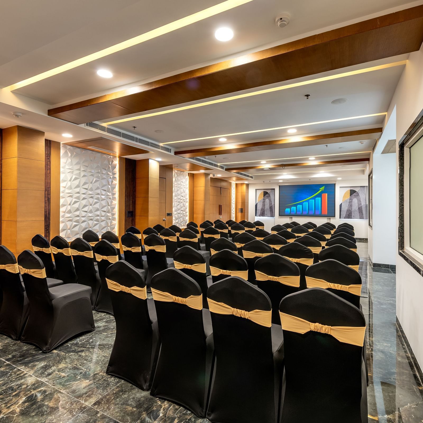 Banquet room with rows of black chairs facing a presentation area, adjacent to large glass windows.
