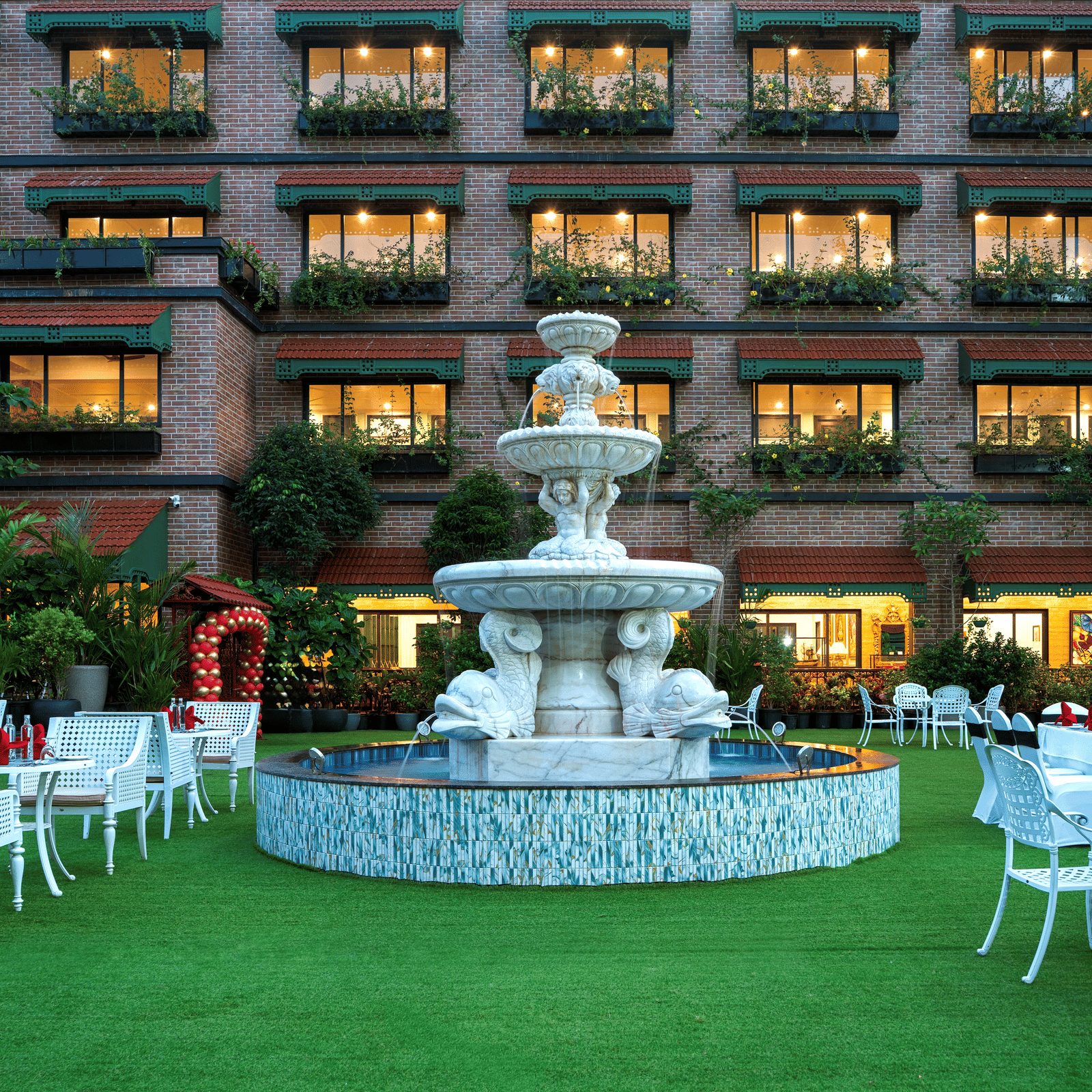 Outdoor dining area on a manicured lawn next to MAYFAIR Bay Resort, Paradeep's facade, set up with white tables and a fountain in the middle.