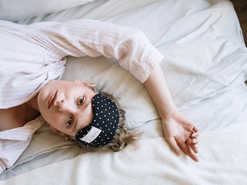 A woman lying awake on white bedding with a polka-dot sleep mask pushed up onto her forehead