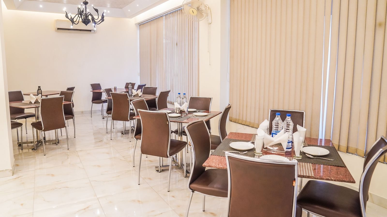 An elegant dining area with multiple tables and chairs is illuminated by a modern chandelier and wall lights - Asian Suites Cyber Park, Gurgaon