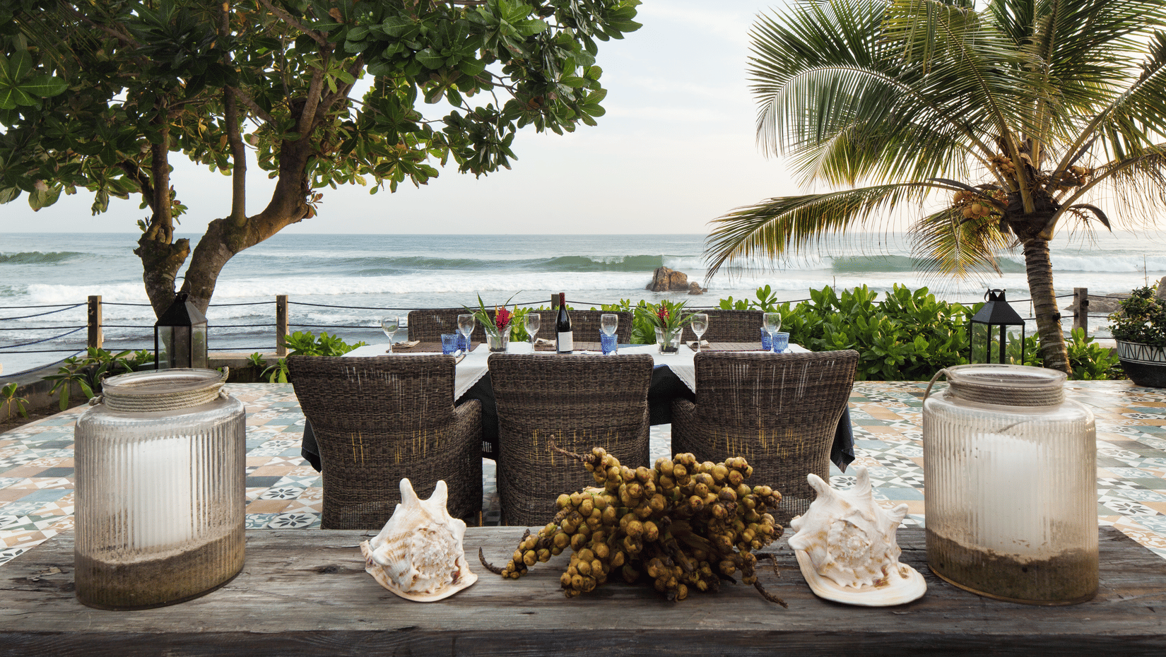 Dining tables with wicker chairs and glass lanterns on the deck, facing the surf
