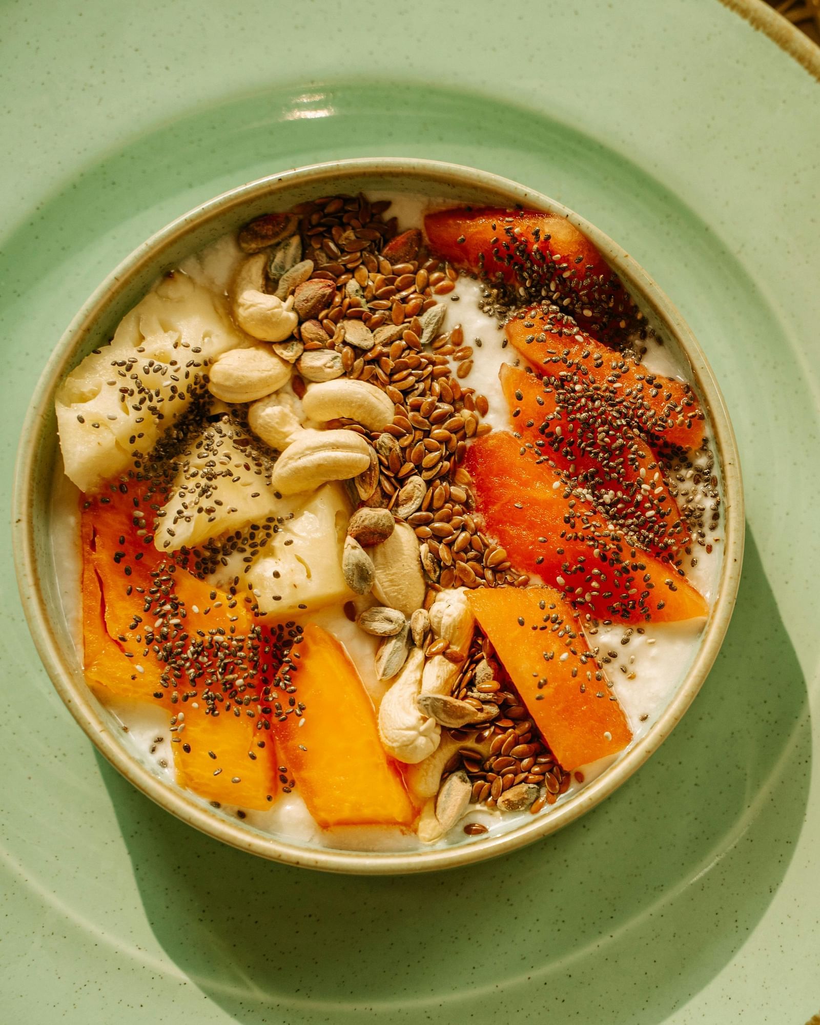 A bowl of fruit topped with seeds, nuts, and yogurt, along with a glass of juice and dessert on the side