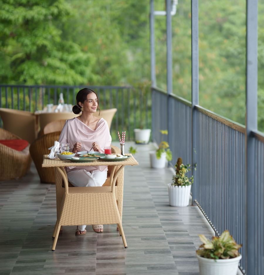 A woman is seated at a table on an outdoor balcony overlooking lush green trees and a scenic landscape, enjoying a meal or drink at Summit Enigma Resort and Spa.