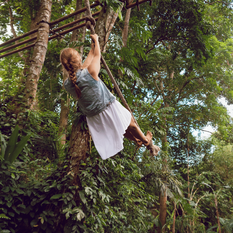 A girl in a frock swinging a tarzan swing, tied to a tree top, in a dense forest.