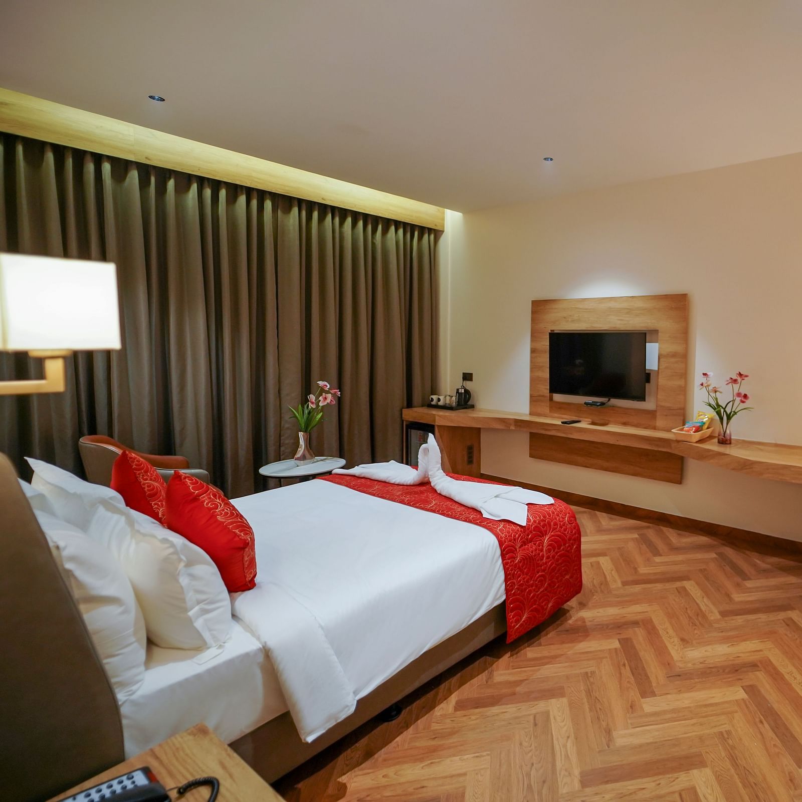 A hotel room features a bed with pillows, a mounted television, a desk with a lamp and chair, curtains, and patterned flooring - Hotel Park Inn, Nellore.