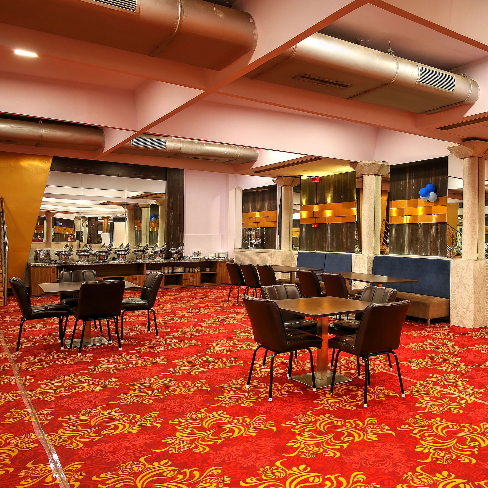 A banquet area with a red patterned carpet, dining tables with chairs, and a bar with blue and gold accents under warm lighting.