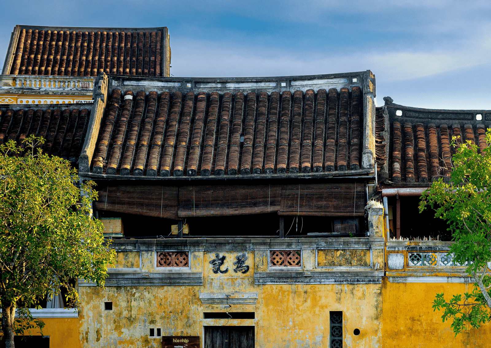 An overview of an old building with people walking in front of it and trees on either side of the building..