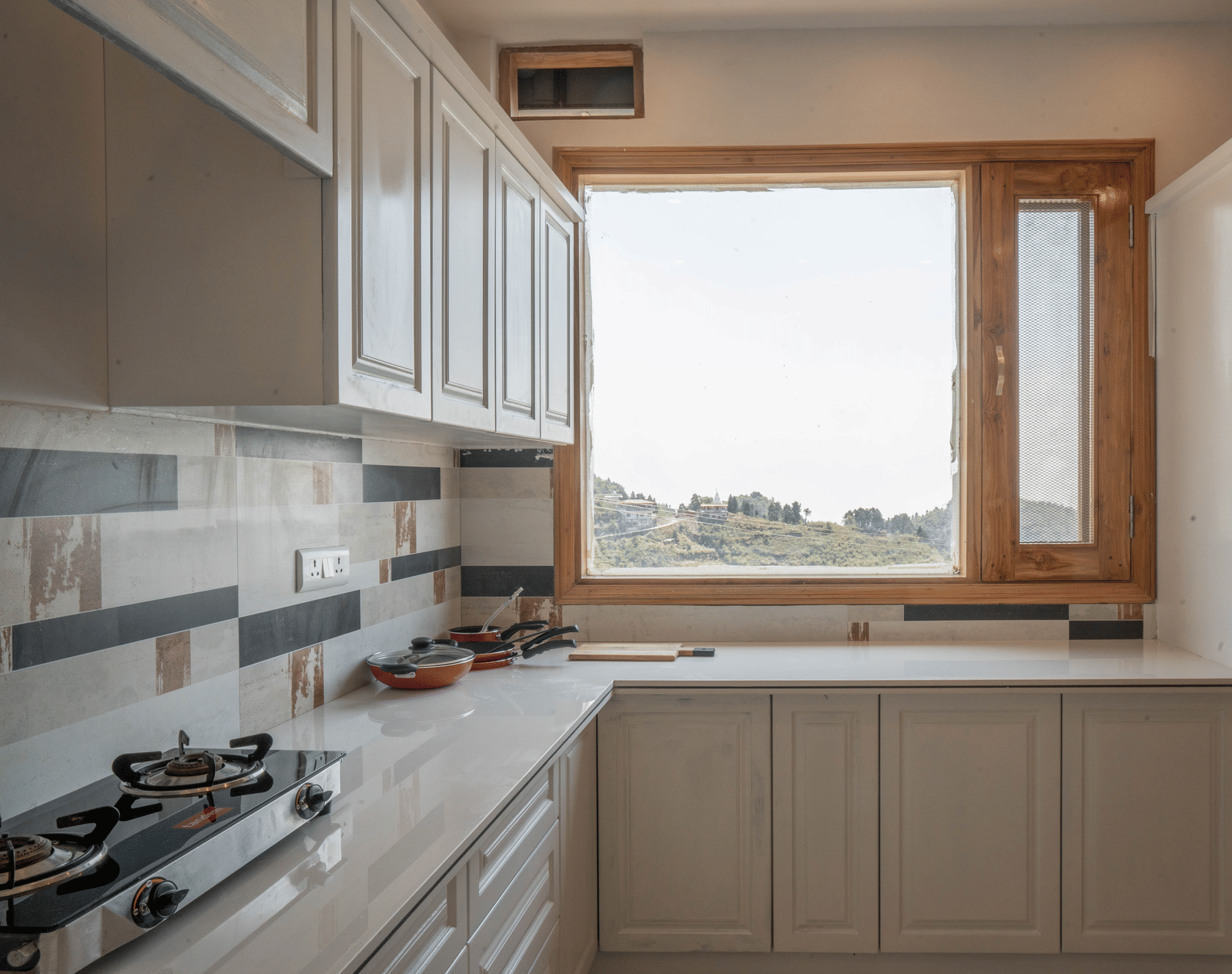 A kitchen at Perfectstayz Value Paradise, Mussoorie, featuring a gas stove, cabinets, and a large window offering a view of the hills.