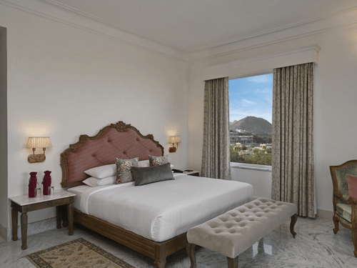 A well-lit Heritage Chamber Suite at Khas Mahal Suites, Udaipur with a large bed, a tufted bench at the foot of the bed, a chair, a nightstand with a lamp, and a window offering a view of an outdoor landscape.