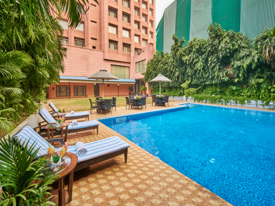 An outdoor swimming pool during the day with blue water, lounge chairs, and a multi-story building in the background | HHI Varanasi