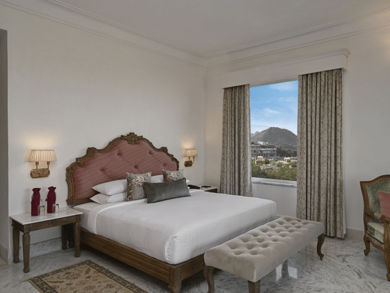 A well-lit Heritage Chamber Suite at Khas Mahal Suites, Udaipur with a large bed, a tufted bench at the foot of the bed, a chair, a nightstand with a lamp, and a window offering a view of an outdoor landscape.