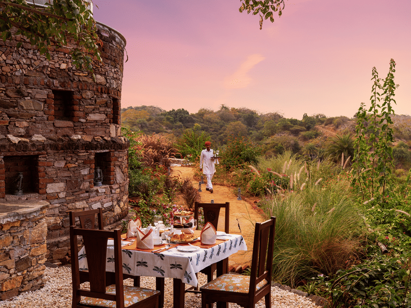 A table for 4 set up next to a stone building with forest cover in the backdrop at Chunda Shikar Oudi, Udaipur