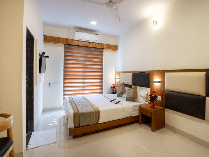 A bedroom at Home2 India with a double bed, pillows, bedside table, wall lights, window with blinds, wall paneling, and a door visible on one side