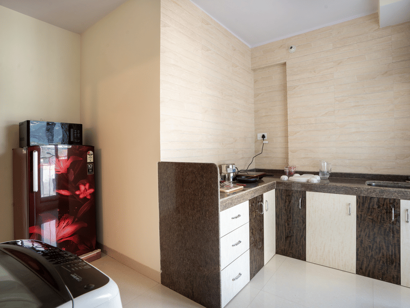 A kitchen area at Home2 India with countertop, sink, cabinets, drawers, tiled walls, a refrigerator, and part of a washing machine visible in the foreground.