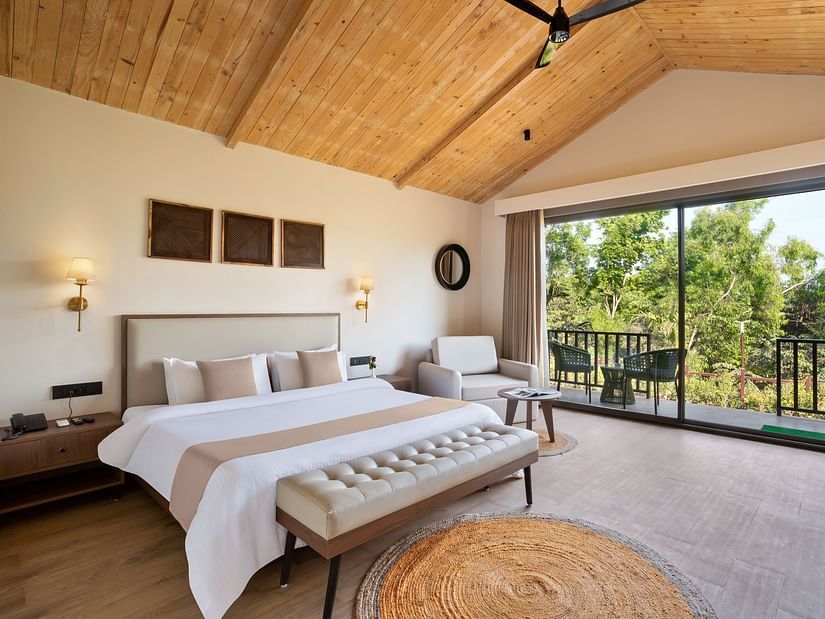 An image of a premium cottage featuring a king size bed, a bedside table and lamp on either side, a sofa chair, and a spacious balcony attached to the room at Stone Woods Premier Resort, Gokarna