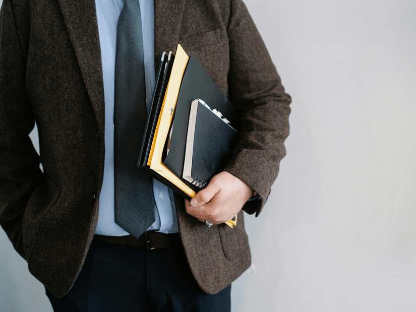 A person dressed in a brown blazer, light blue shirt, and dark tie holding several folders and a notebook under their arm, with one hand in their pocket, against a plain light background | Retail Summit Mumbai