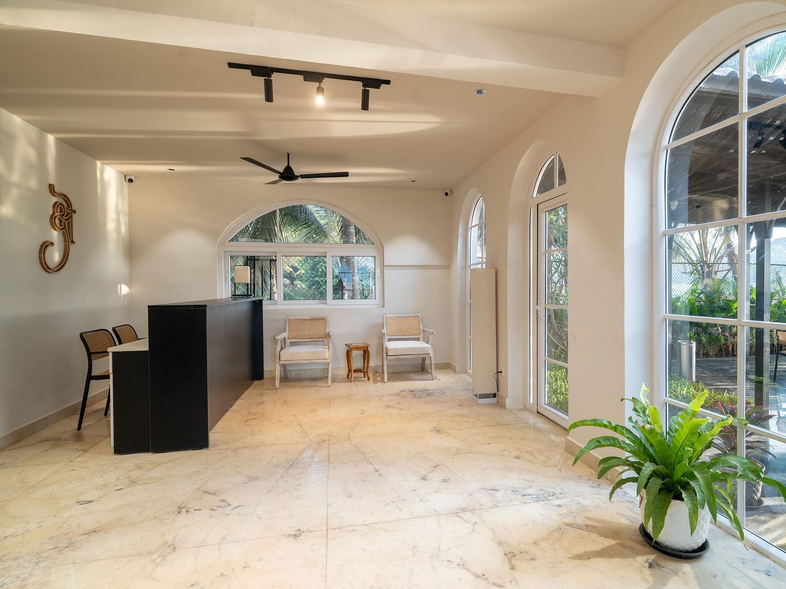 A bright reception space with a polished floor, leading towards arched windows showing greenery outside. A black front desk sits to the left, and a potted plant at Kssemya