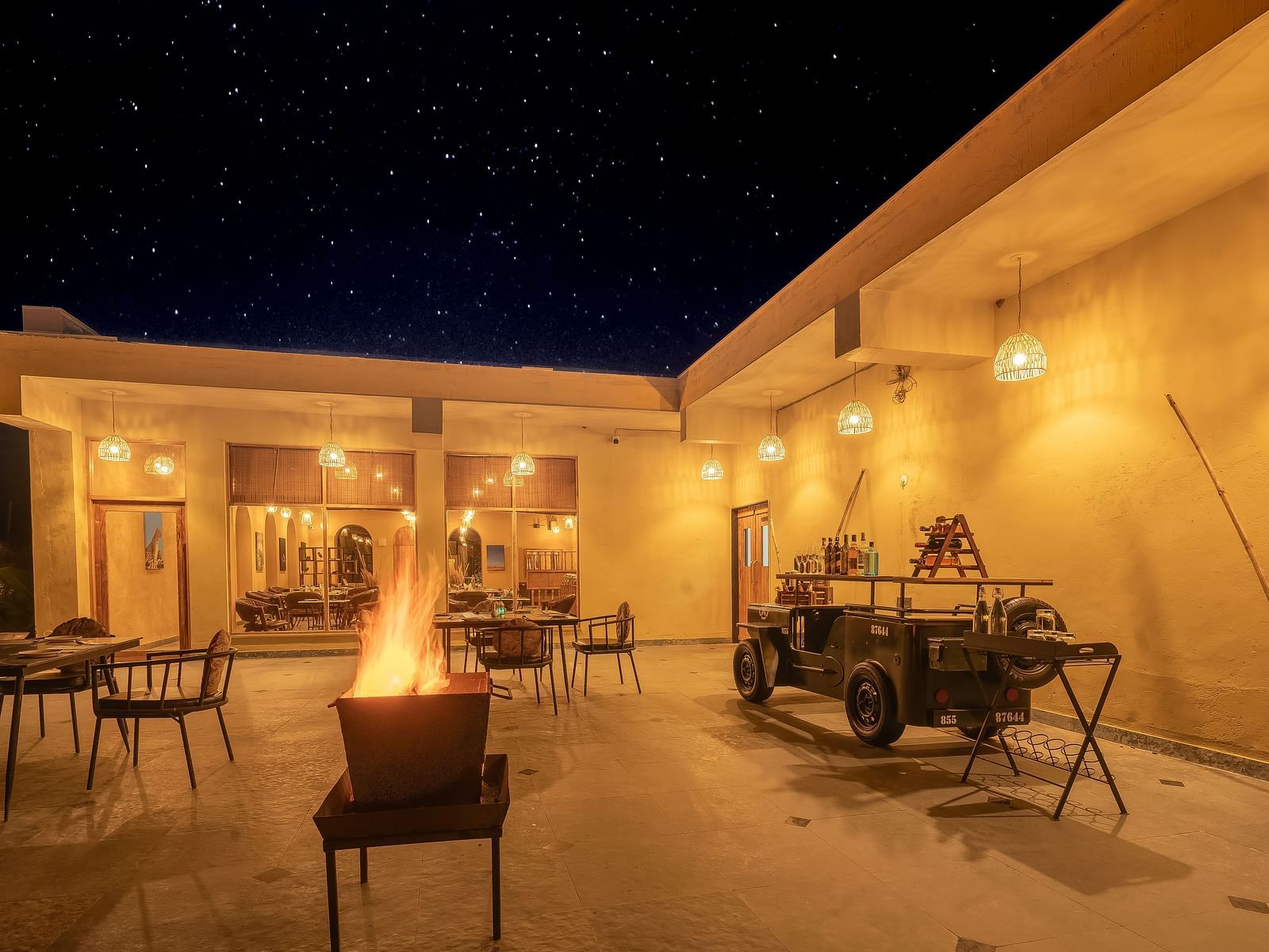An image of open air Rosette Restaurant featuring several sets of table and chairs, a jeep without roof meant for decoration, and a pot where fire is buring at Kurja Jawai