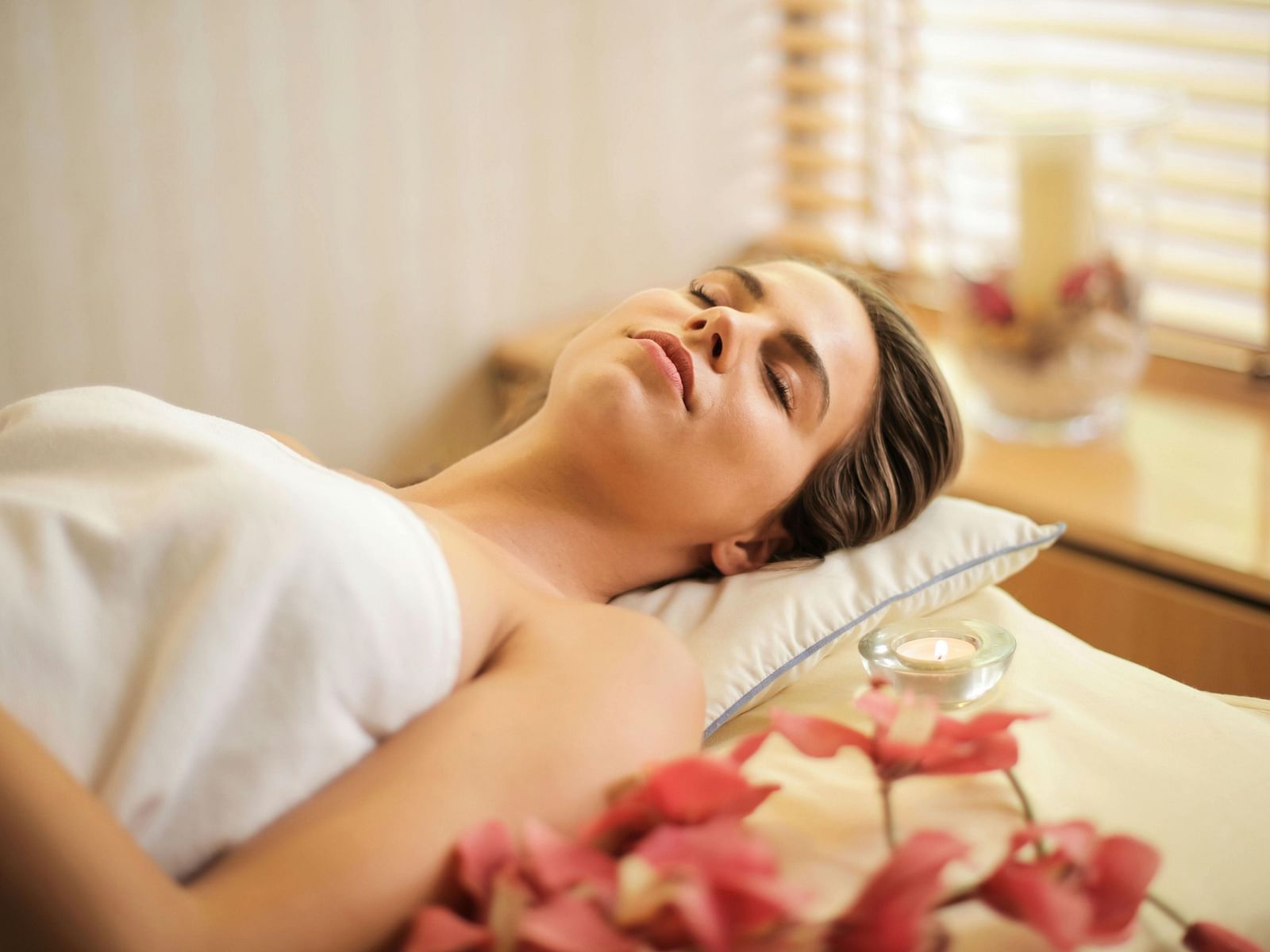A woman lay on a spa bed, wrapped in a white towel with candles and flowers visible nearby.