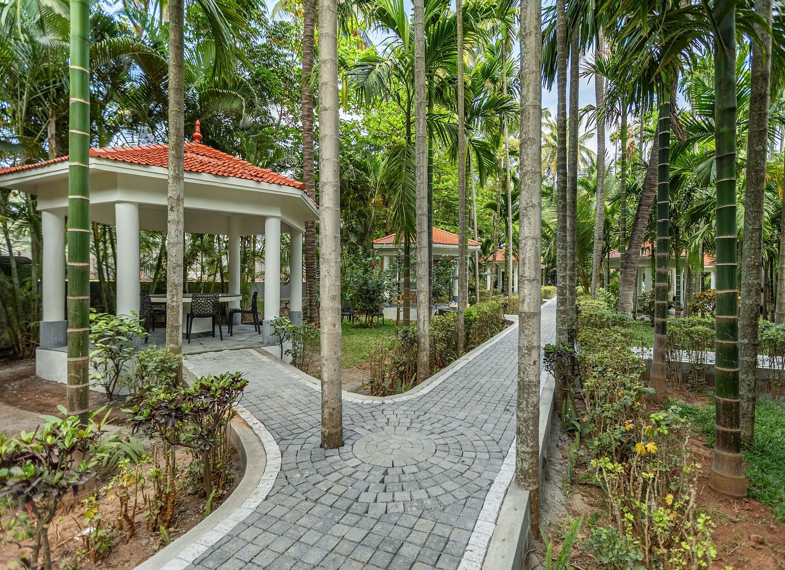 A white gazebo with an orange roof and a paved pathway winding through palm trees and greenery - Mango Hill Resort, Coimbatore