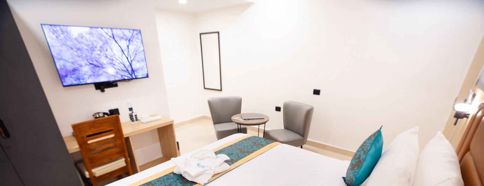 View of the room from beside the headboard, showing a neatly made bed facing a wall-mounted TV and a study table with a chair below at Sunrise Business Hotel, Hyderabad.