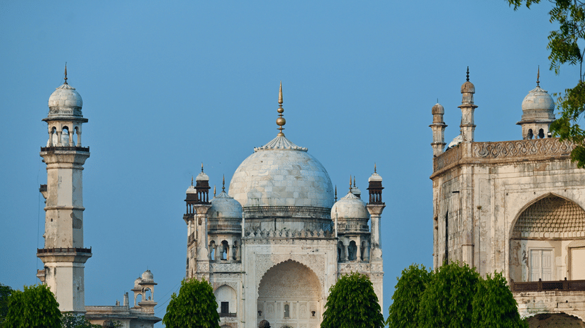 A wide shot of a large white mausoleum (Bibi Ka Maqbara) with domes and minarets, surrounded by green trees against a clear blue sky