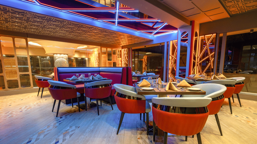 A dining area with tables set with chairs on a light-coloured floor, under a wooden roof structure, illuminated with blue lights at night | Pride Elite Chhatrapati Sambhajinagar