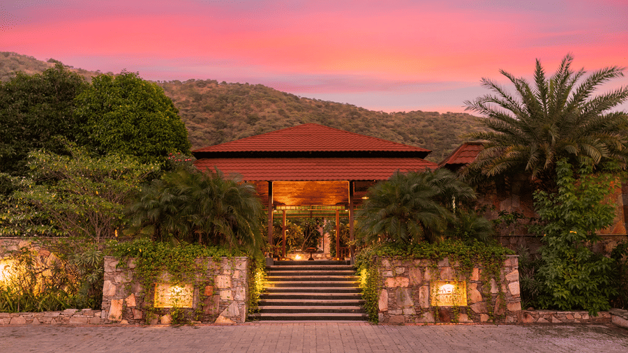 The resort entrance with red roofs and stone walls illuminated against a pink twilight sky | Anandam Jungle Retreat Resort, Udaipur