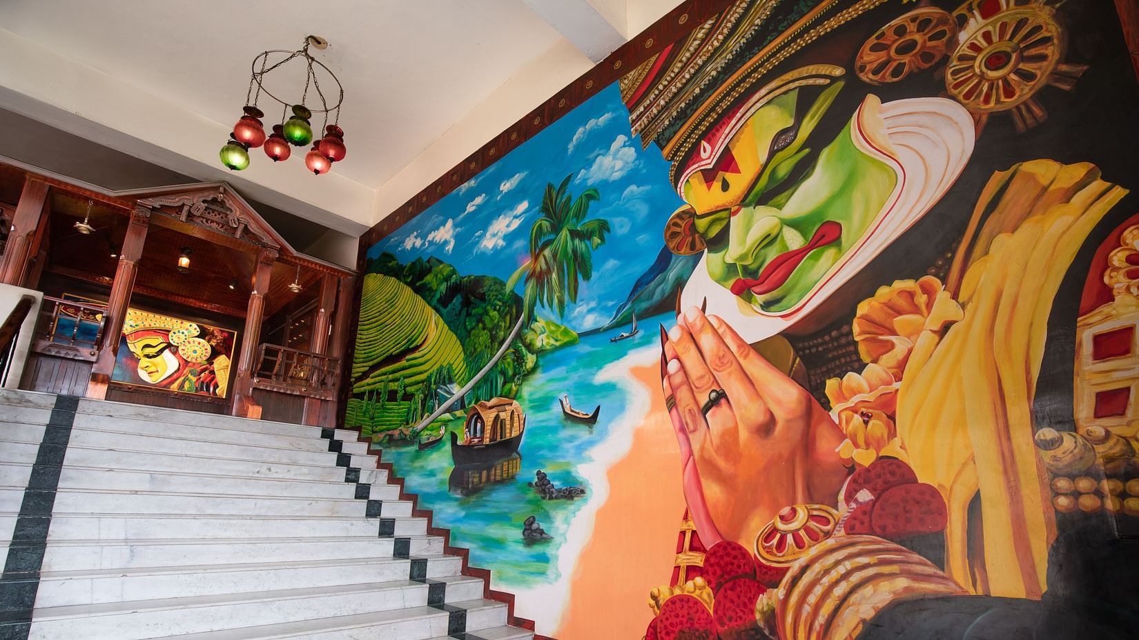 A vibrant mural featuring a traditional Kathakali dancer, tropical scenery, and boats decorates the wall beside a wide staircase at Jenneys Residency with ornate wooden carvings and a hanging chandelier