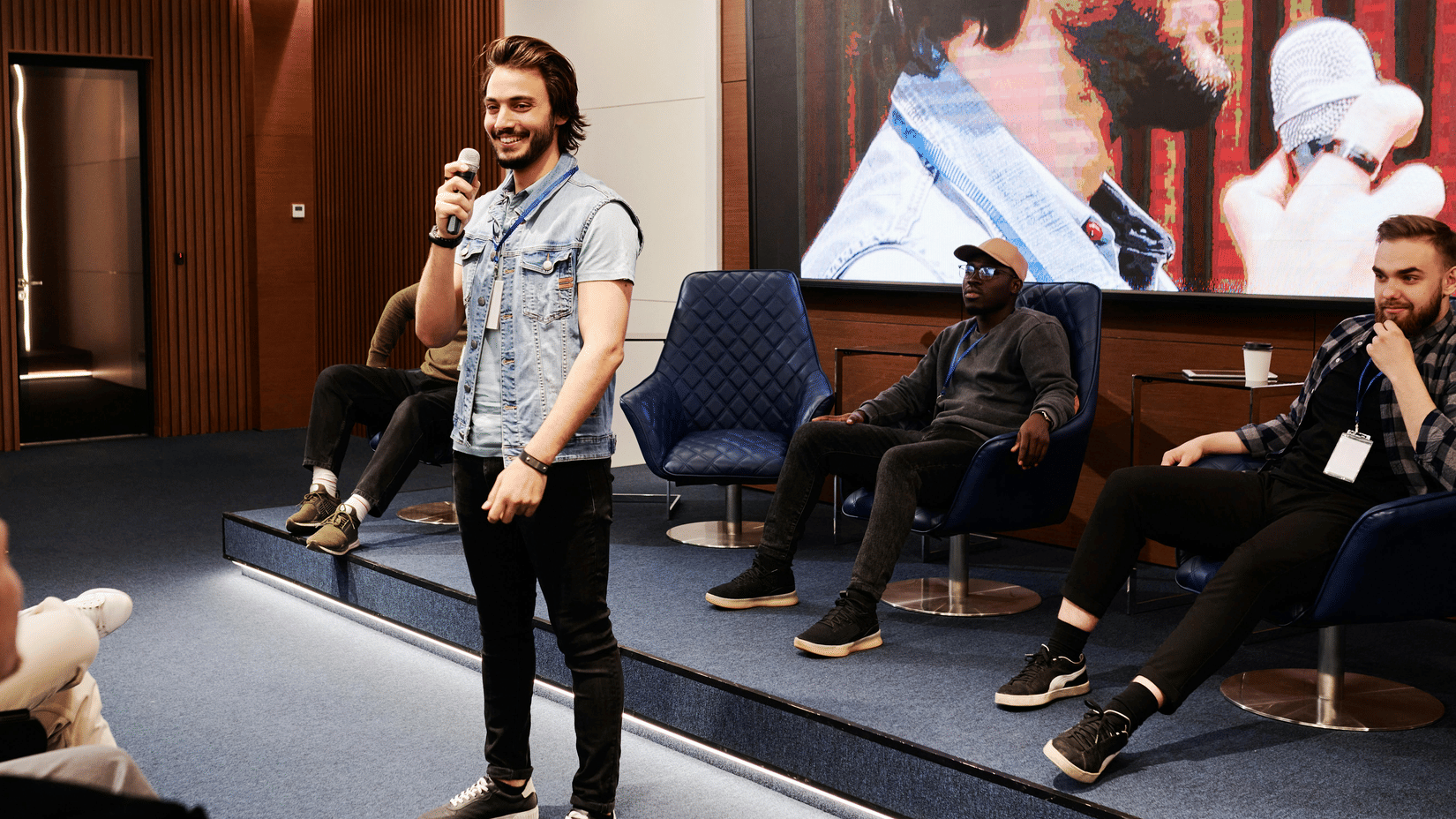 A male speaker wearing a light-coloured shirt and dark trousers stands on a stage with a microphone addressing an audience.