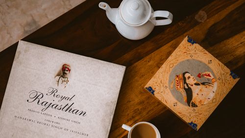 A peaceful coffee setting features a cup, teapot, heritage book titled “Royal Rajasthan,” and intricate artwork, evoking refined leisure at Colonel’s Retreat, Defence Colony.