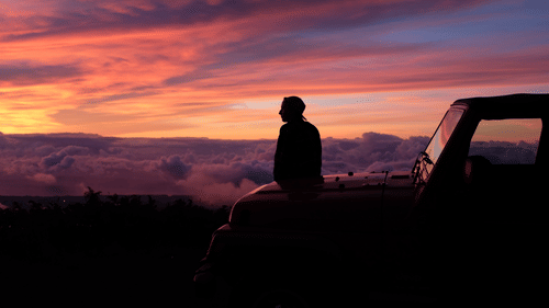a person sitting on the hood of a car looking at the different hues in the sky