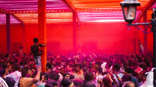 A large group gathered under a covered space during Holi celebrations, surrounded by pink and red coloured powder.