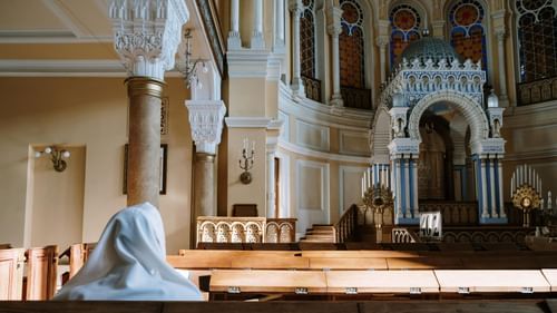 A person seated on a bench wearing a shawl, looking at the pulpit of the Mattancherry Synagogue.