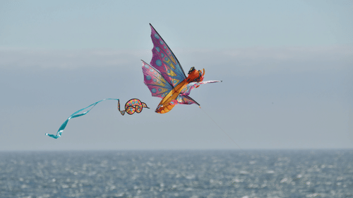 A colourful butterfly-shaped kite flying high above the shimmering surface of the ocean on a bright day.