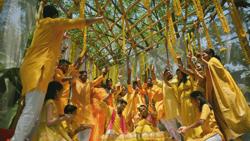 A couple sitting together in a decorated stool meant for Haldi ceremony. The couple is surrounded by other people who are also wearing yellow coloured dresses. 
