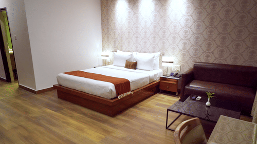Bedroom of Junior Suite at Sarovar Portico, Bhubaneswar, showcasing the plush king size bed placed under ample overhead lighting, beside a seating area, and over wooden flooring.