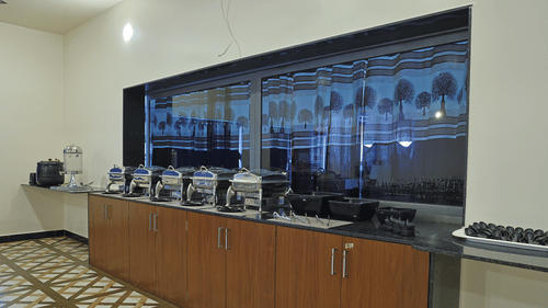 A buffet counter at the VITS Select Diamond Resort, Sasan Gir restaurant. The counter has several steel serving dishes and bowls, with a large window and patterned curtains behind them.