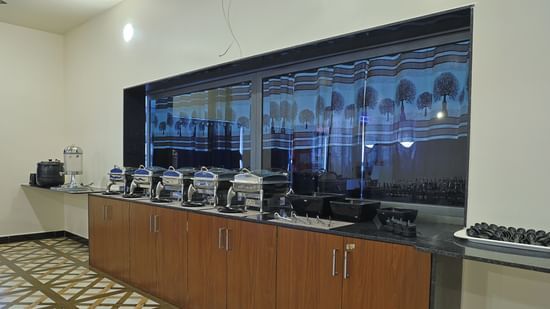 A buffet counter at the VITS Select Diamond Resort, Sasan Gir restaurant. The counter has several steel serving dishes and bowls, with a large window and patterned curtains behind them.