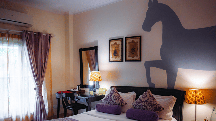 An elegant bedroom features rich fabrics, warm lighting, and a distinctive horse silhouette wall art, highlighting artistic flair and comfort at Colonel’s Retreat in Delhi.