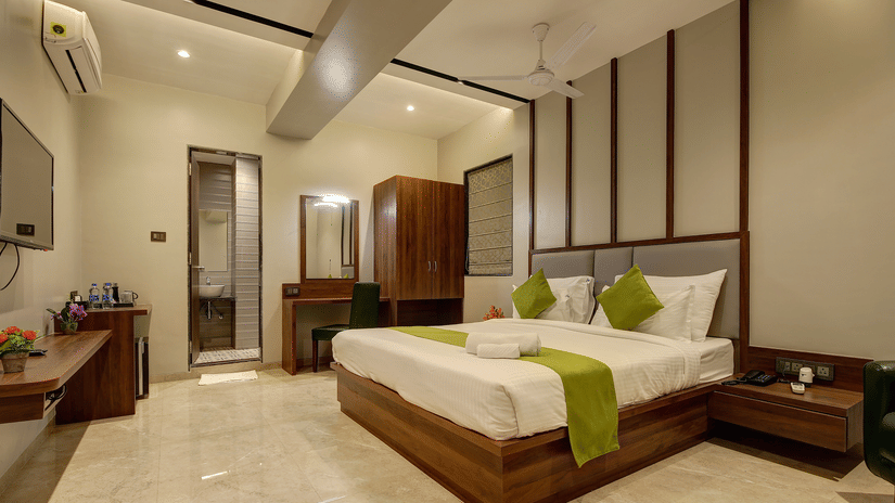 A spacious, well-lit hotel room featuring a wooden double bed with white linens and green accents, a vanity desk, wardrobe, wall-mounted TV, seating area, and an attached bathroom - Magnus Fortune House, Magarpatta