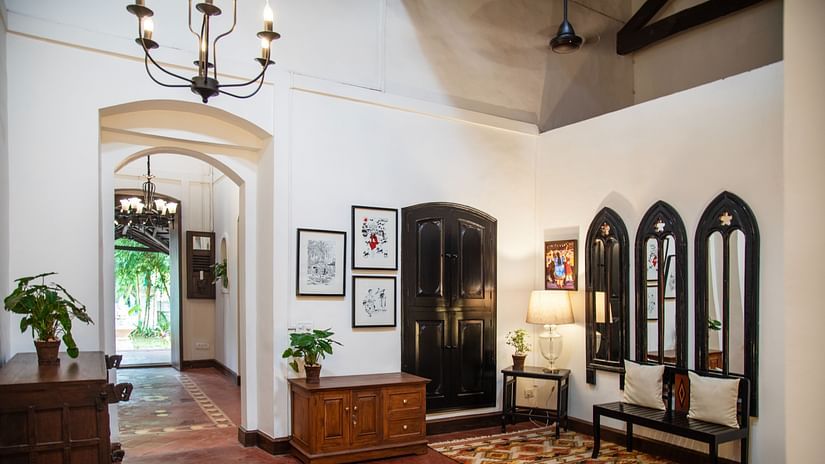 An image of a huge lobby where various decorative items are placed on top of the cabinets and shelves, there is another table with lamp and three vintage style window near it - Lamrin Ucassaim Hotel, Goa