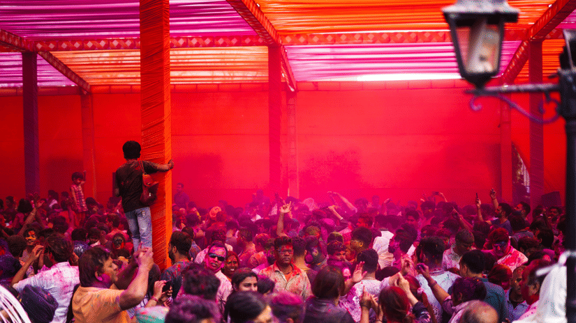 A large group gathered under a covered space during Holi celebrations, surrounded by pink and red coloured powder.