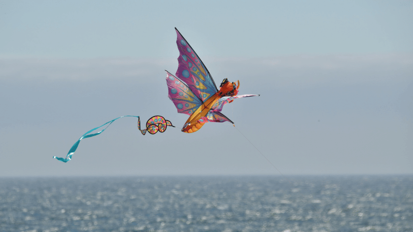 A colourful butterfly-shaped kite flying high above the shimmering surface of the ocean on a bright day.