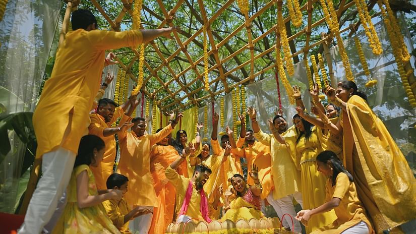 A couple sitting together in a decorated stool meant for Haldi ceremony. The couple is surrounded by other people who are also wearing yellow coloured dresses. 