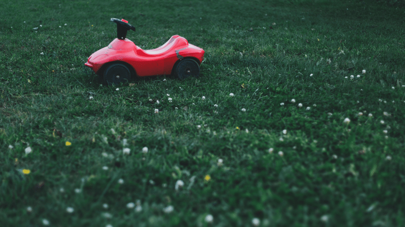 A small, red plastic ride-on toy car is sitting on a grassy lawn with white specks of clover or daisies.