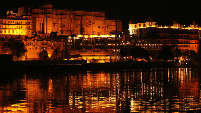 Beautifully illuminated heritage buildings of Gangaur Ghat reflected in the waters of Lake Pichola at night.