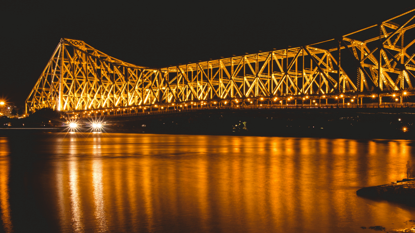 The massive steel cantilever Howrah Bridge illuminated with golden lights, reflecting beautifully on the Hooghly River at night.